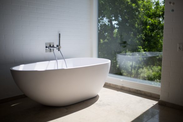 Solitary freestanding bathtub next to a full glass wall window with green foliage on the outside.